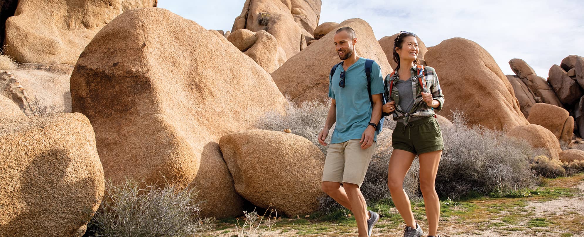 Man and woman wearing backpacks hike along a trail surrounded by large boulders during their WorldMark by Wyndham vacation.