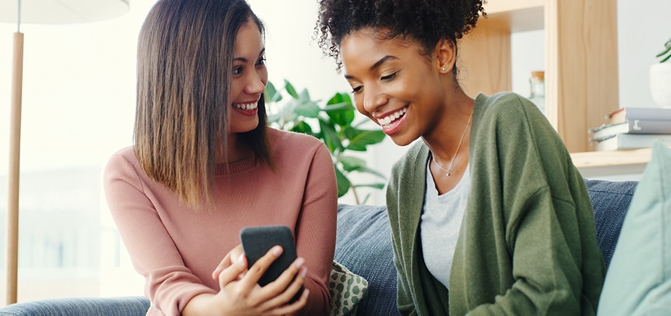 A woman holding a cellphone smiles at her friend who is looking down at the phone. 