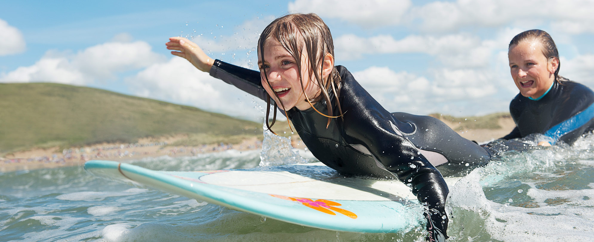 A young kid wearing a wetsuit paddles on a surfboard as a woman pushes the board into a wave.