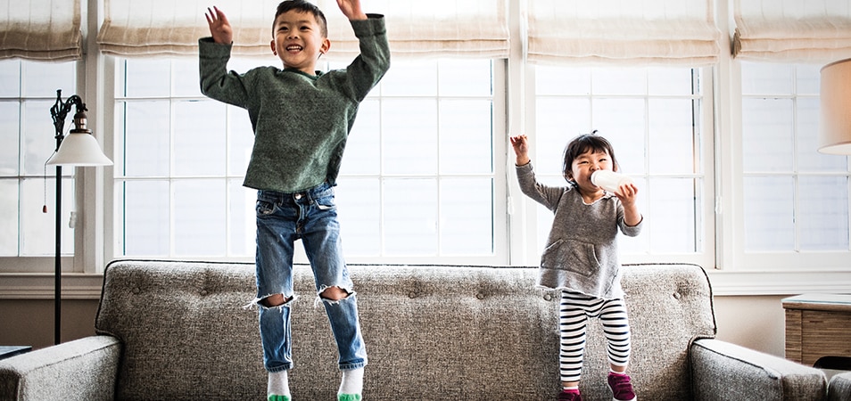 Two young kids jump on a couch with their arms above their heads.