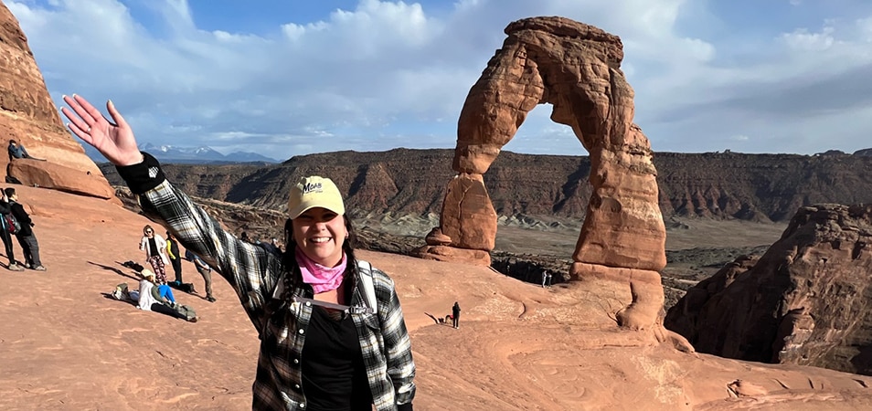 A woman in hiking gear holds her arm up while posing in front of Delicate Arch at Arches National Park in Moab, Utah. 