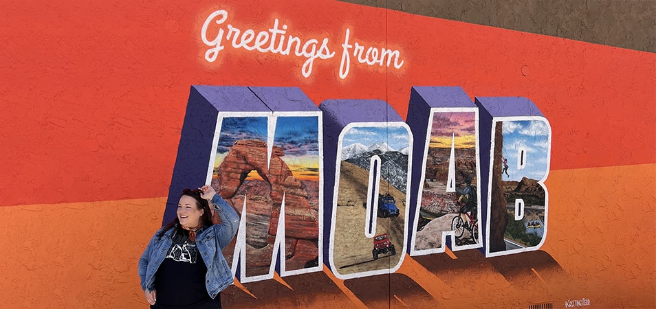A woman looks off to the left while posing in front of a larger mural that says Greeting from Moab while visiting Moab, Utah.