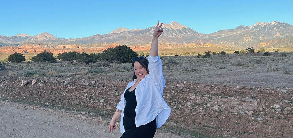 A woman holds a peace sign up in the air as she poses in front of red rock mountains on the side of the road while driving out of Moab, Utah.