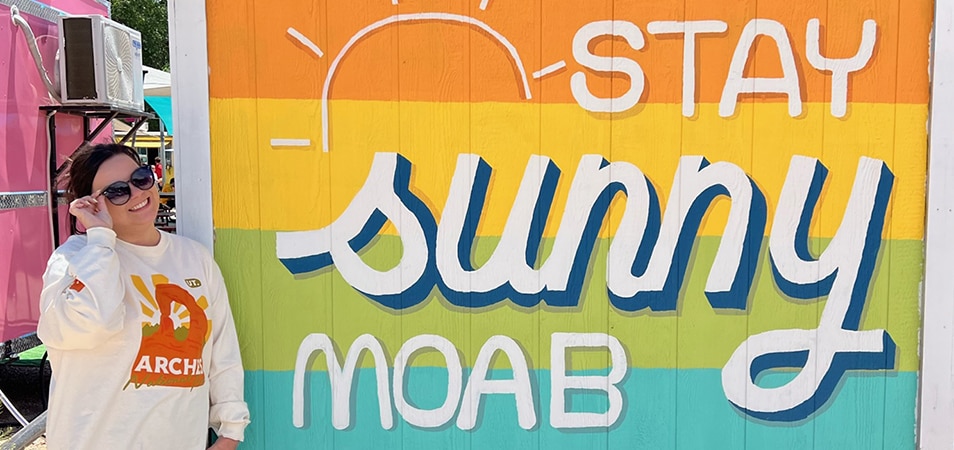 A woman with sunglasses on smiles and stands to the side of a rainbow-colored mural that says Stay Sunny Moab while visiting Moab, Utah.