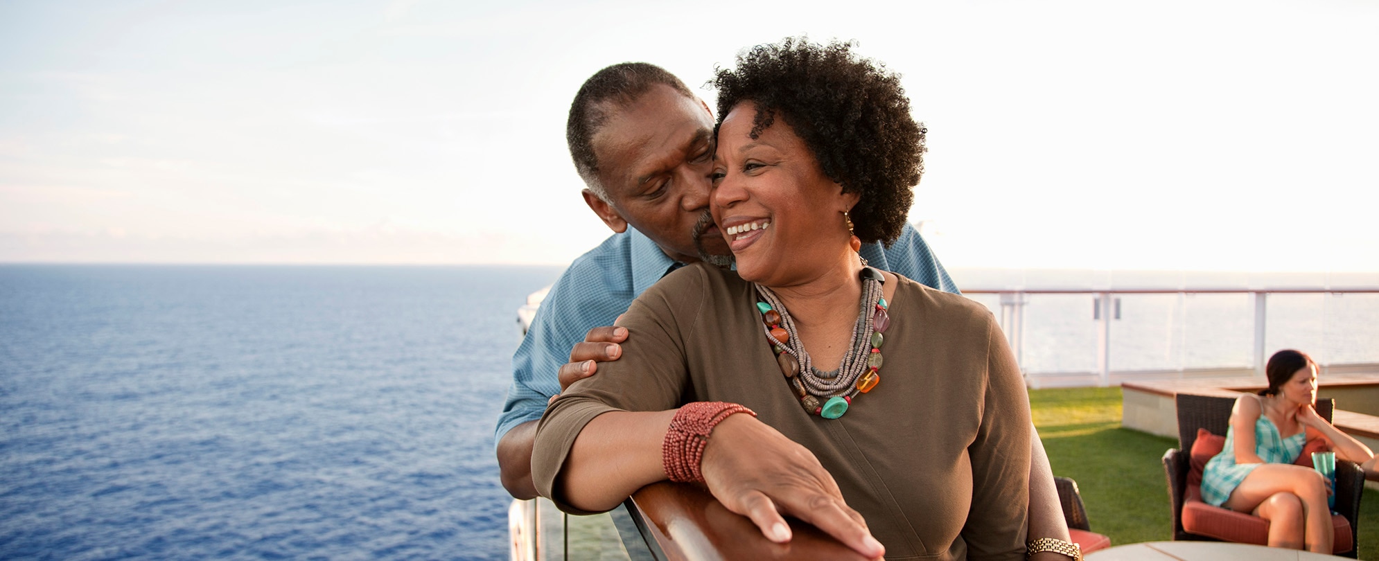 An older man kissing his wife's cheek while smiling on a deck overlooking the water.