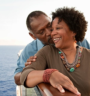 An older man kissing his wife's cheek while smiling on a deck overlooking the water.