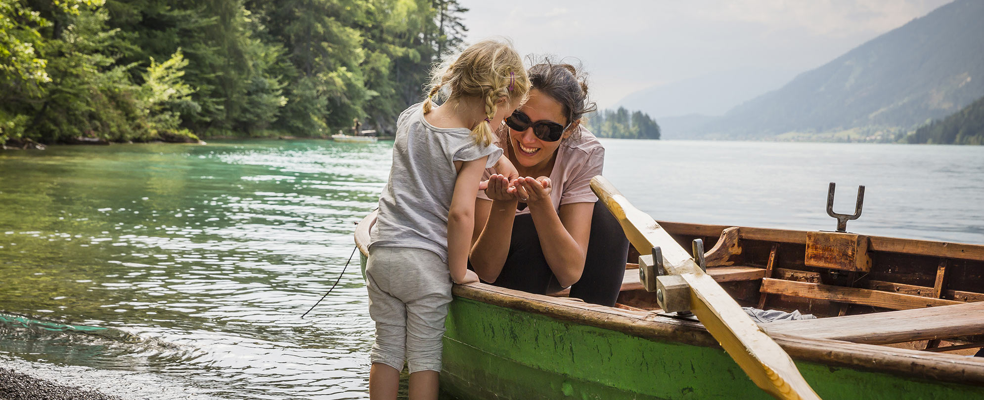 A woman sits in a row boat while docked in the shallow end of a lake holds her hands out for a little girl facing her to grab something from them.