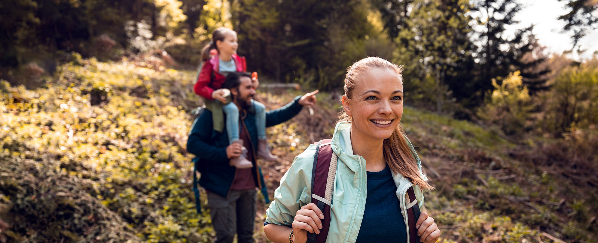 Woman smiles while hiking down a hill with a man walking behind her carrying a young girl on his shoulders and pointing to something out in the distance.