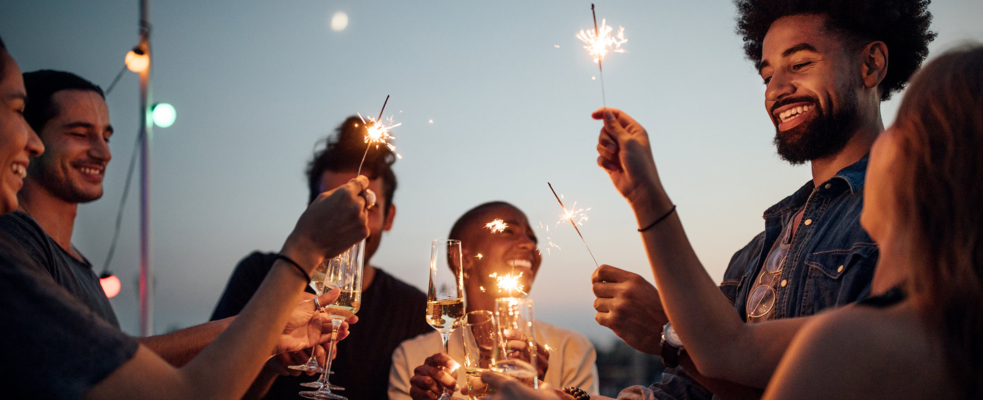A group of friends stand together as they toast glasses of champagne and raise sparklers in celebration.