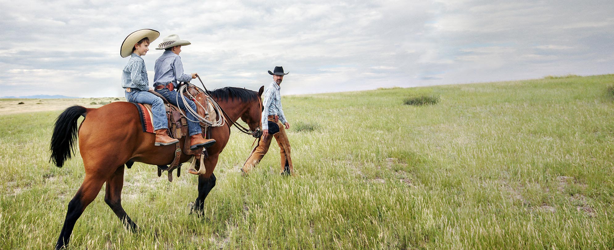 Two boys dressed in in jeans, flannel shirts, boots, and cowboy hats ride on a brown horse as a man in cowboy attire walks in front of them through  a grassy field.