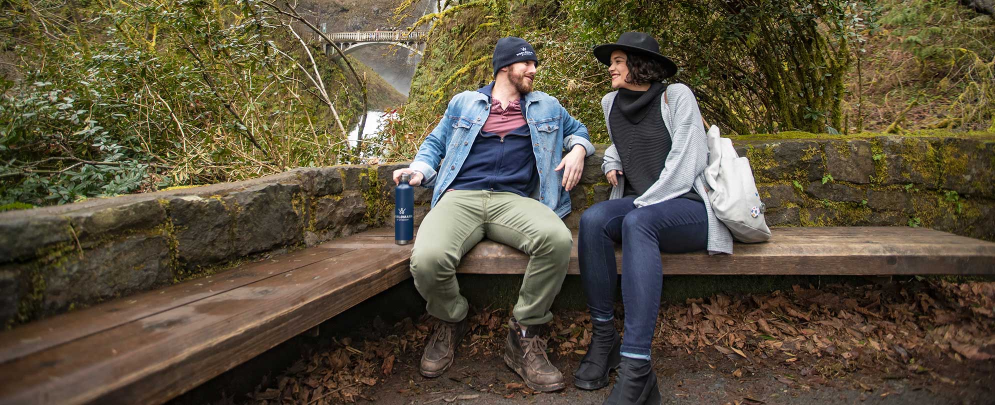 A couple, sitting next to each other on a wooden bench in the woods, conversating. The man is wearing a WorldMark by Wyndham beanie and is holding a WorldMark by Wyndham bottle. 