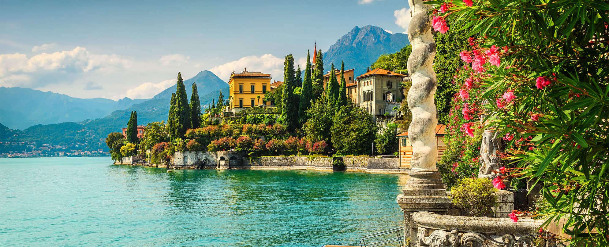 A bright blue lake surrounded by mountains and tall green trees at Villa Monastero on Lake Como in Verenna, Italy.