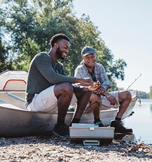 A dad and his adult son smile as they sit on a canoe by the shore of a lake, putting bait on a fishing line. 