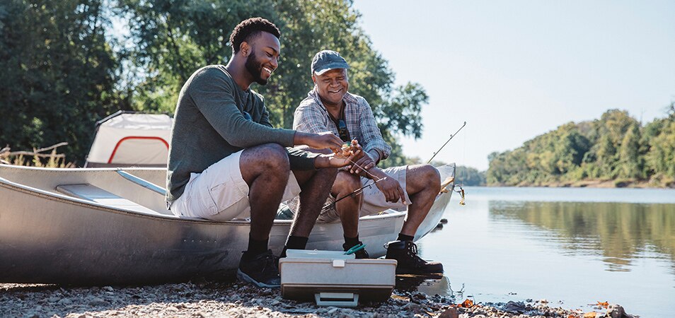 A dad and his adult son smile as they sit on a canoe by the shore of a lake, putting bait on a fishing line. 