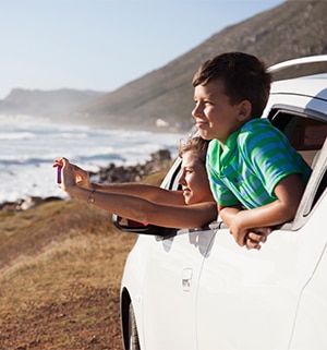 Two children lean out of car window while taking a photograph of beautiful beach scenery.