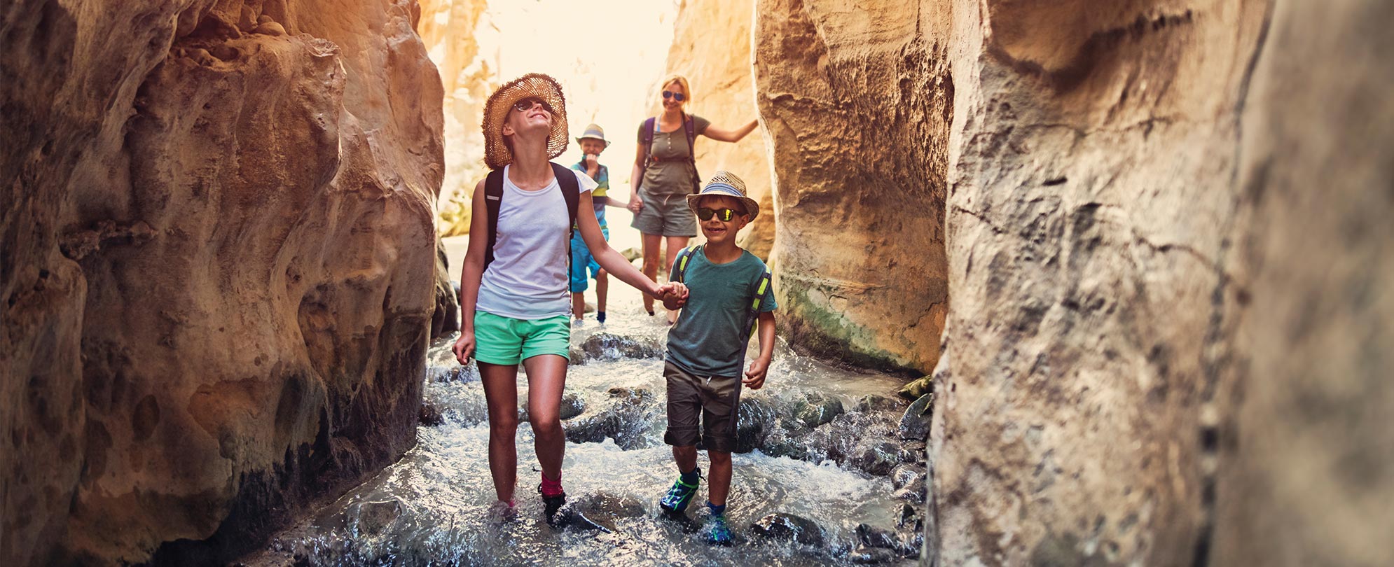 Two moms and two children holding hands while hiking down a stream, between canyons.