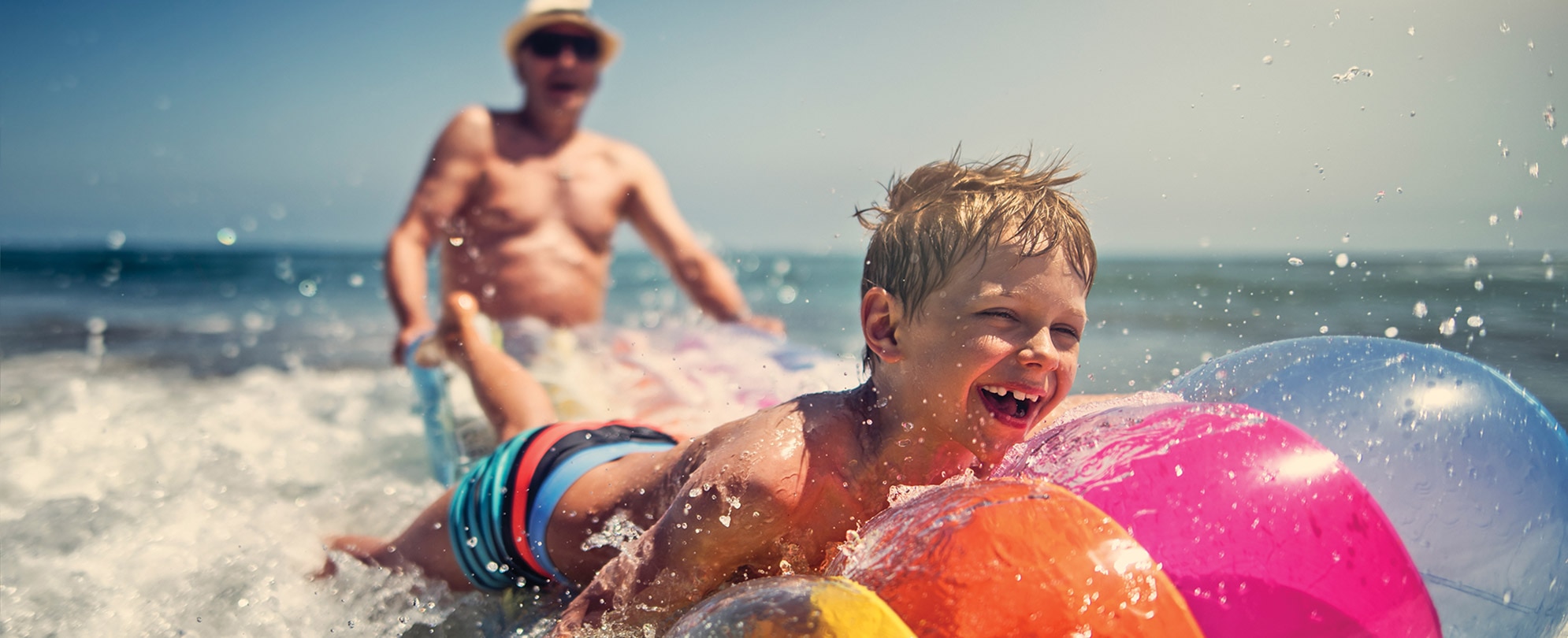 A smiling young boy splashes through the water on a colorful inflatable raft with his grandpa standing behind him.