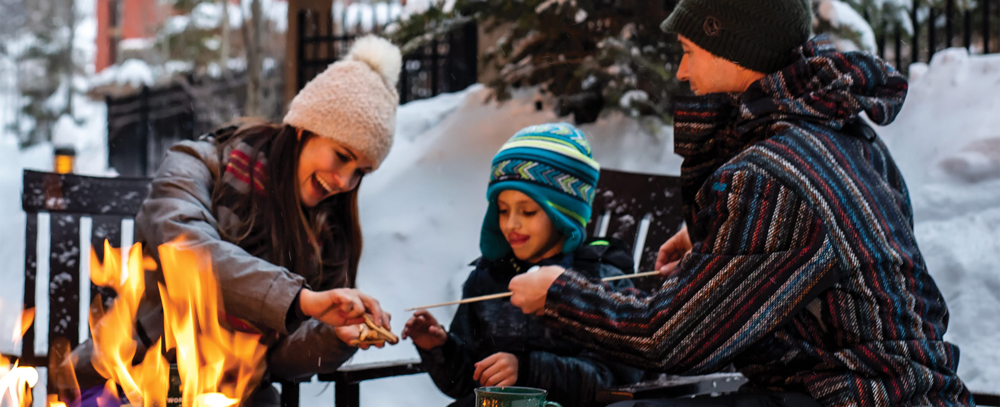 A mom, dad, and young son wearing winter clothes roast marshmallows around a fire at a WorldMark by Wyndham timeshare resort.