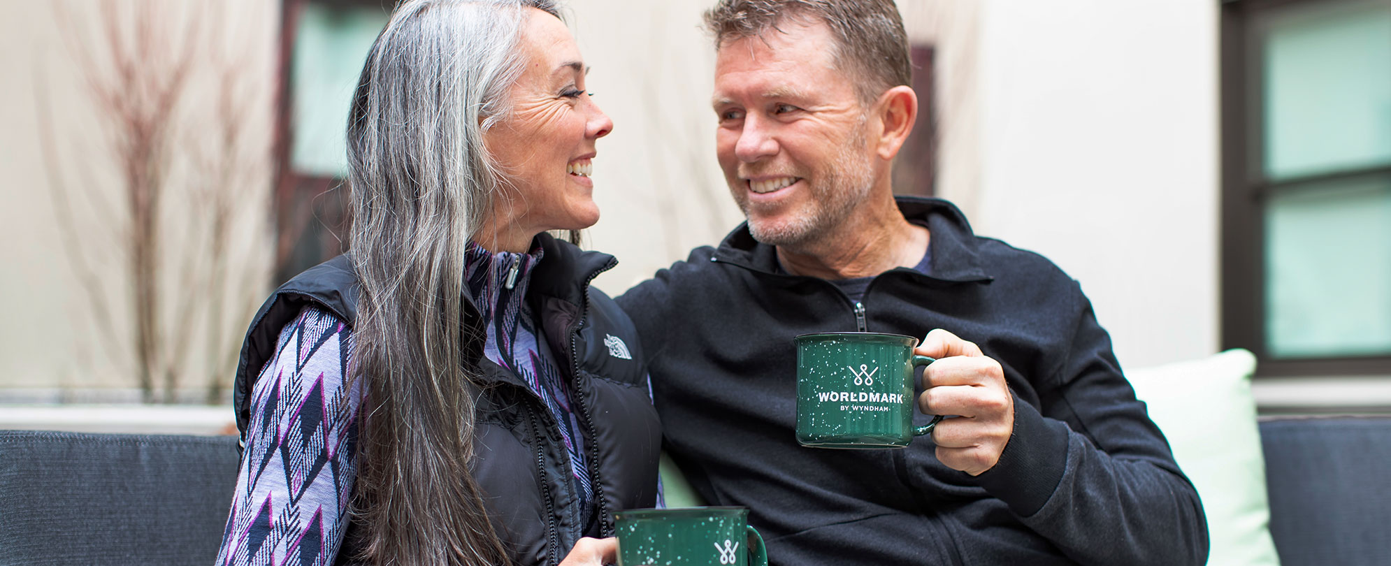 Older man and woman smiling at each other holding coffee mugs with the WorldMark by Wyndham logo.