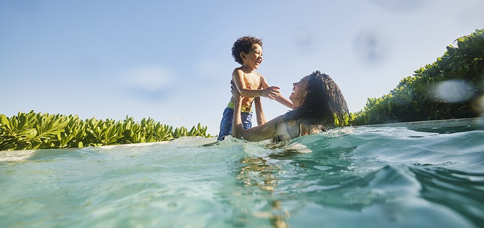 A woman holds a little boy up out of the water as they both laugh while swimming in the ocean.