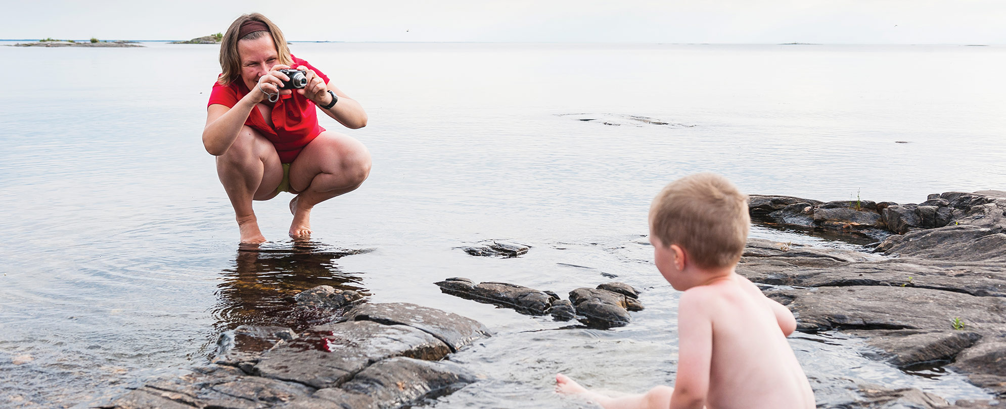 A mom bends down to take a photo of her young son on a rocky shoreline.