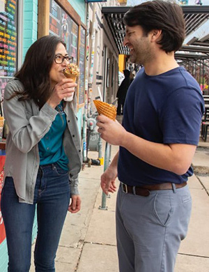 A smiling man and woman enjoy ice cream cones while standing in front of the store window.