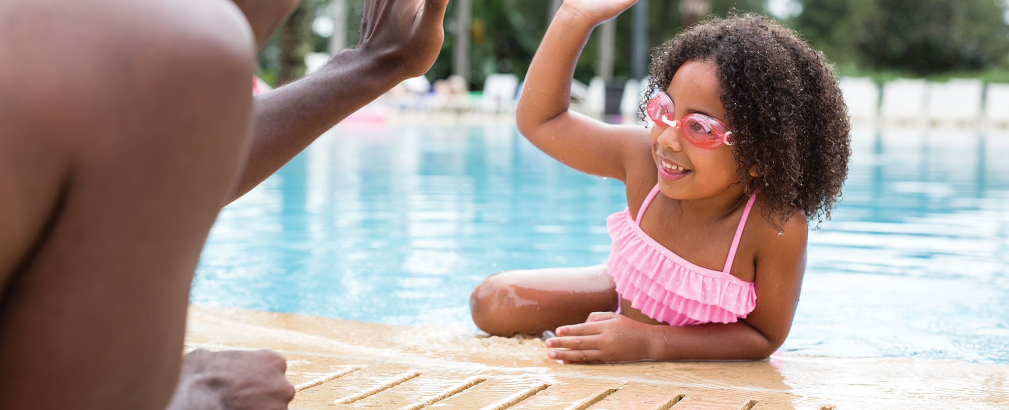 A young girl wearing goggles for swimming leans out of a WorldMark by Wyndham resort pool, high-fiving her parent.