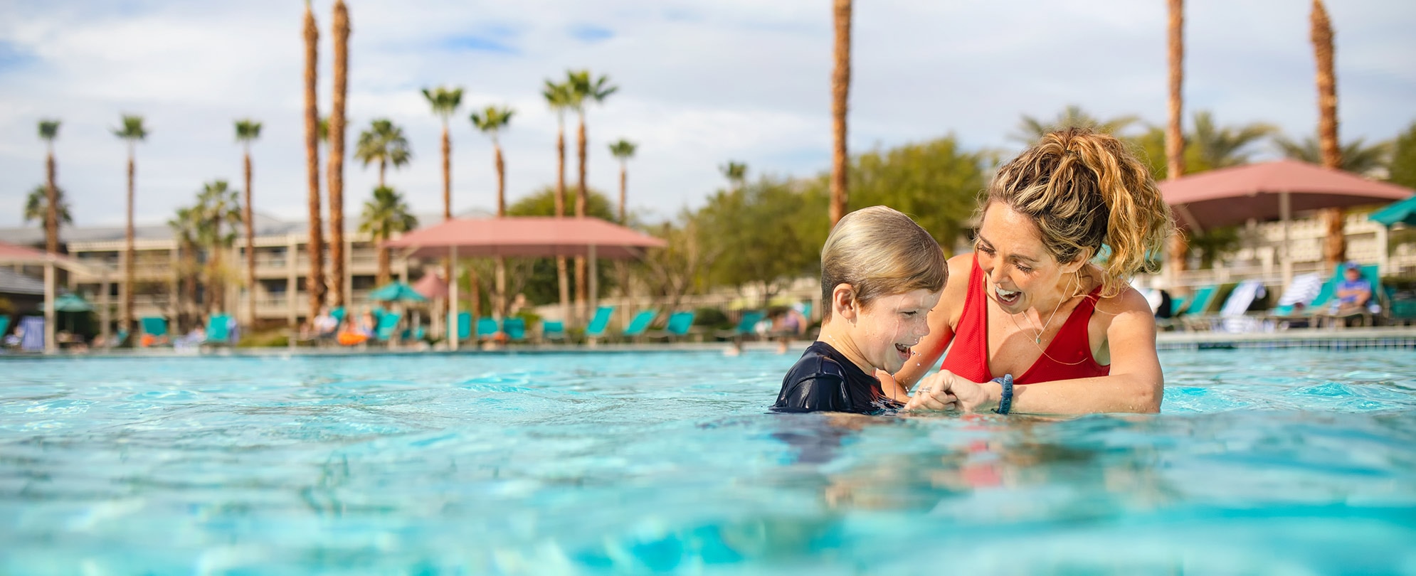 Mom in a red swimsuit and her son smile and splash in a pool on their WorldMark by Wyndham timeshare vacation.