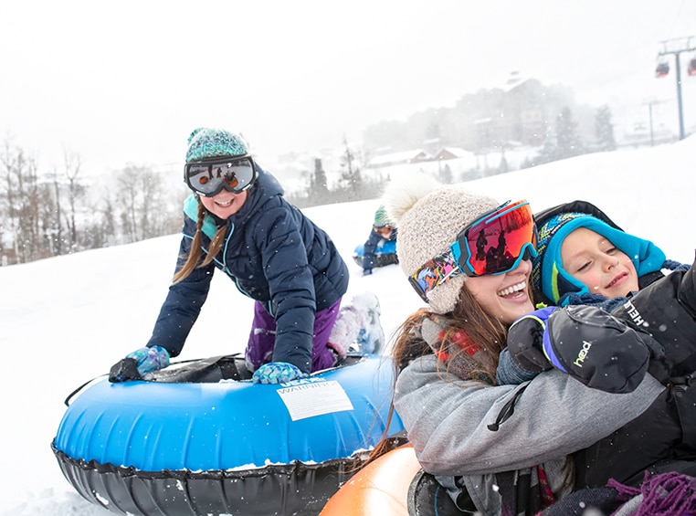 A smiling family in coats, hats, and ski goggles ride snow tubes during their WorldMark by Wyndham winter vacation.